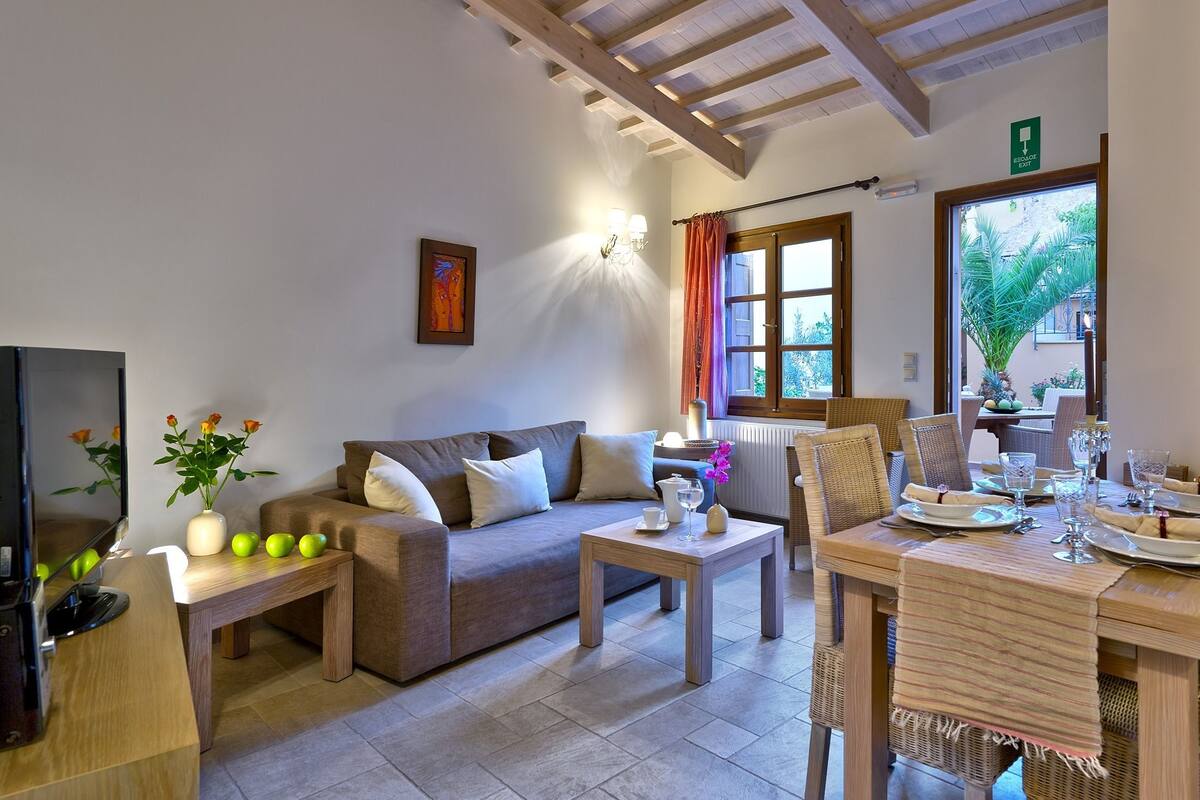 A cozy living room with a sofa, coffee table, and dining area with wicker chairs and a table set for two.