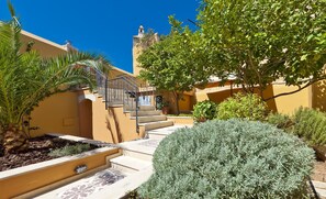 Um pátio com uma escadaria que leva a um edifício, rodeado por vegetação e um céu azul límpido.