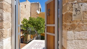 A narrow alleyway with a wooden door and a signboard, leading to a building with a stone facade and a tree in the background. - Kores Boutique Houses (Chania)