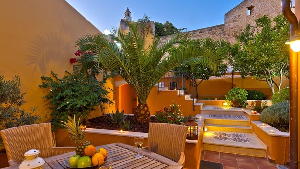 An outdoor patio with wicker chairs, a table with fruit and drinks, and a staircase leading to a building with a bell tower. - Kores Boutique Houses (Chania)