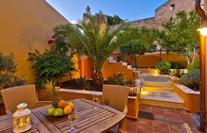 An outdoor patio with wicker chairs, a table with fruit and drinks, and a staircase leading to a building with a bell tower. - Kores Boutique Houses (Chania)