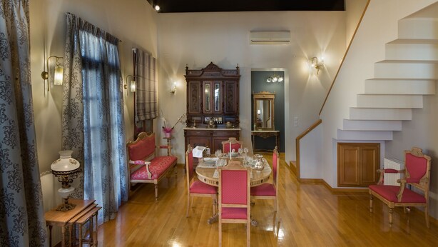 Un coin repas avec une table en bois dressée pour quatre personnes, entourée de chaises à revêtement rose. On aperçoit à l’arrière-plan une grande armoire en bois surmontée d’un miroir.