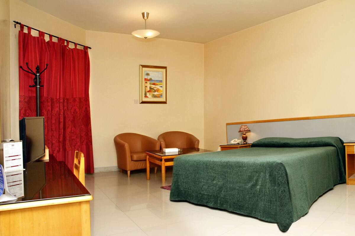 Una habitación de hotel con una cama, dos sillas, un escritorio con una computadora, un teléfono y un cuadro en la pared.