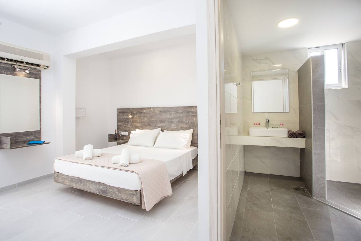A modern bedroom with a large bed, a wooden headboard, a bedside table, a mirror, and a bathroom with a sink and mirror.