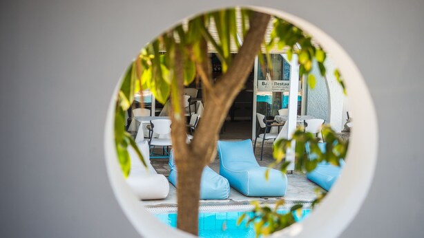 A circular view of a pool area with lounge chairs and a tree, surrounded by a building with a sign reading "Ba. Restcau." - Anika Studios (Rhodes)