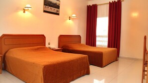 Habitación de hotel con dos camas, una mesita de noche, una lámpara, una ventana con cortinas rojas y un cuadro colgado en la pared.