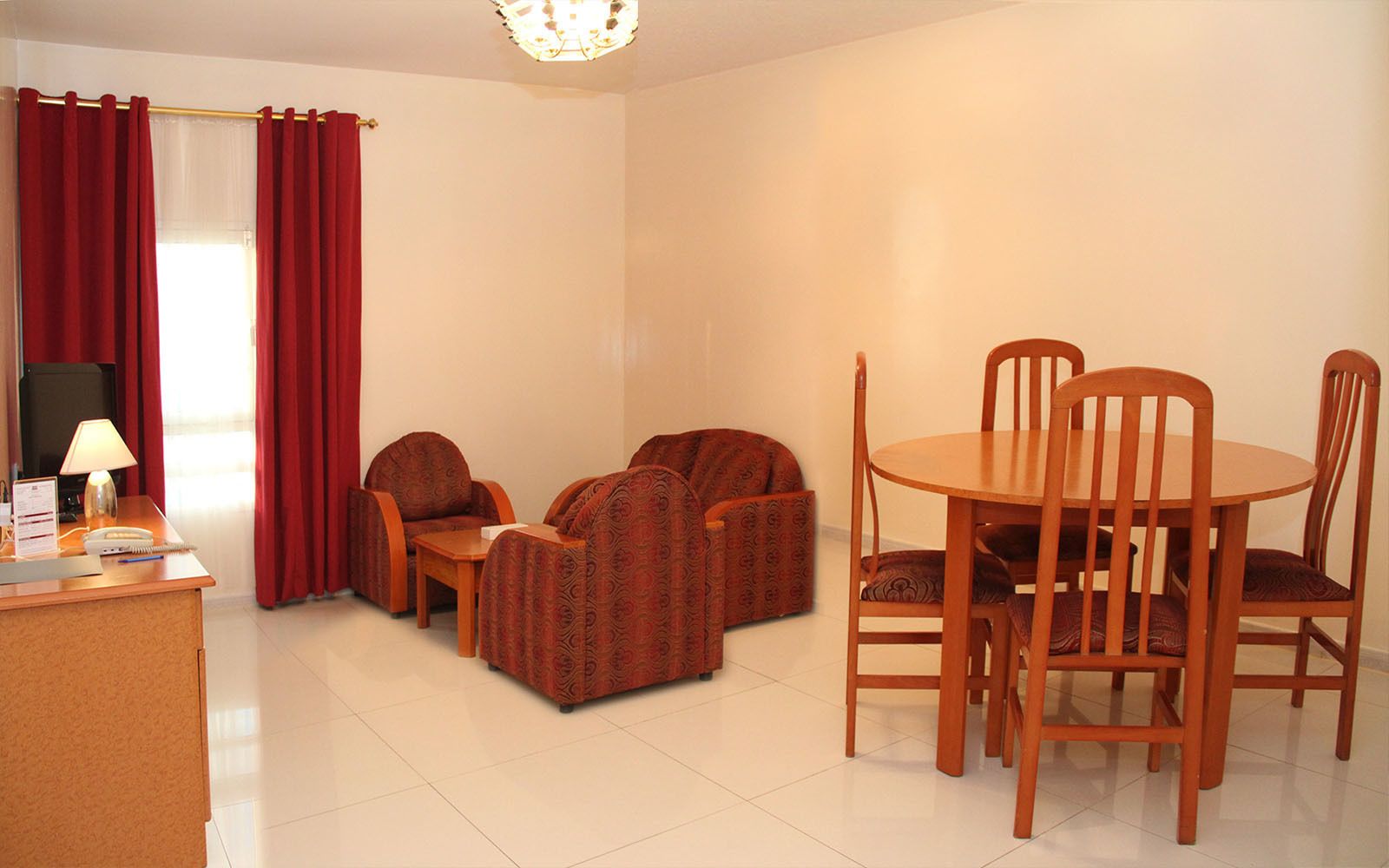 Quarto de hotel com área de refeições, incluindo mesa de madeira e quatro cadeiras, escrivaninha com luminária e telefone, e cortinas vermelhas na janela.