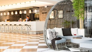 A modern lobby with a checkered floor, a bar area with stools, a seating area with a wicker chair and cushions, and a large potted plant.