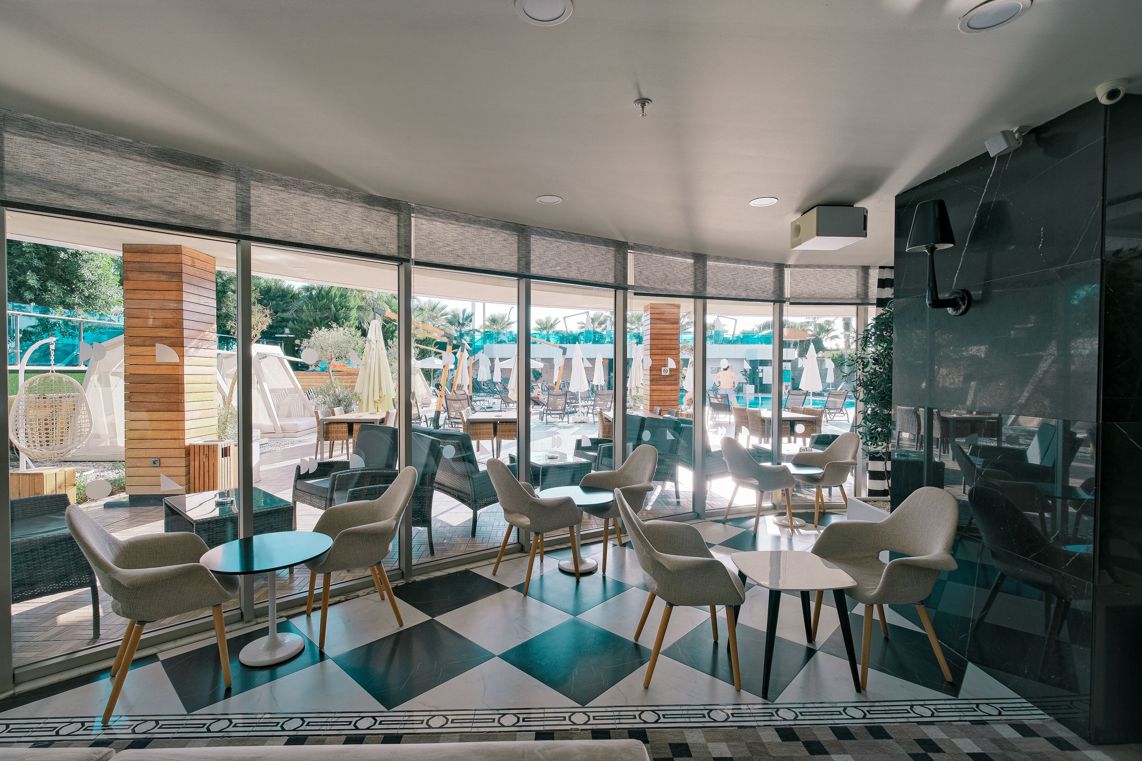 Un área de estar moderna en interiores con paredes de vidrio, suelo a cuadros y vista a una piscina al aire libre.