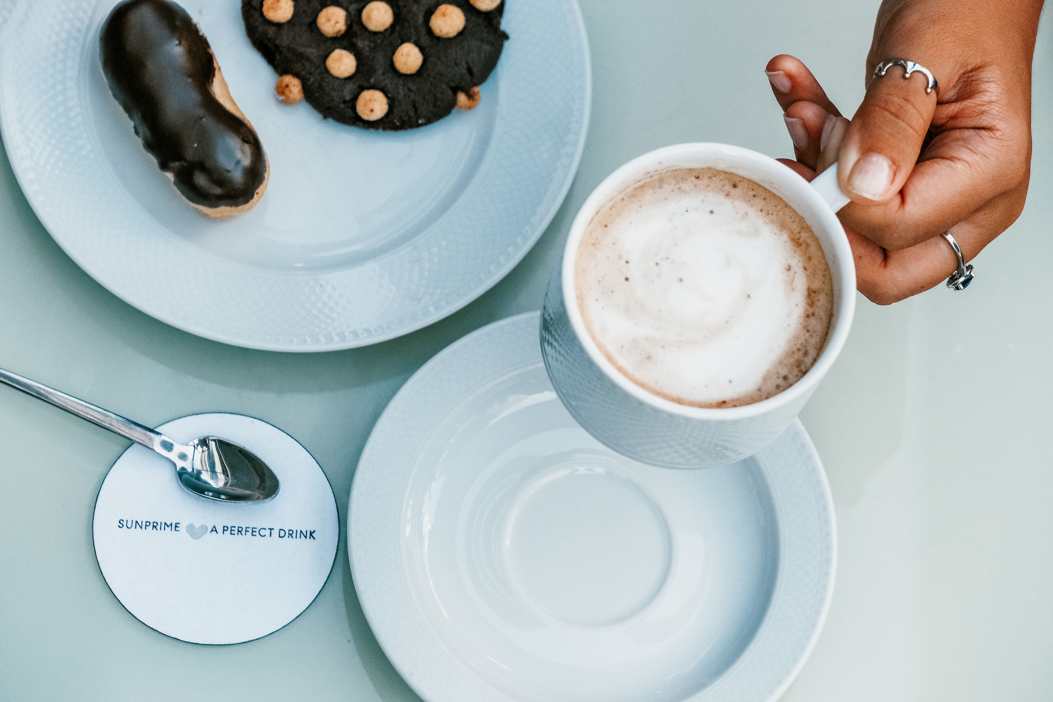 A hand holding a cup of coffee with a saucer, a plate with a cookie, and a spoon on a coaster with text.