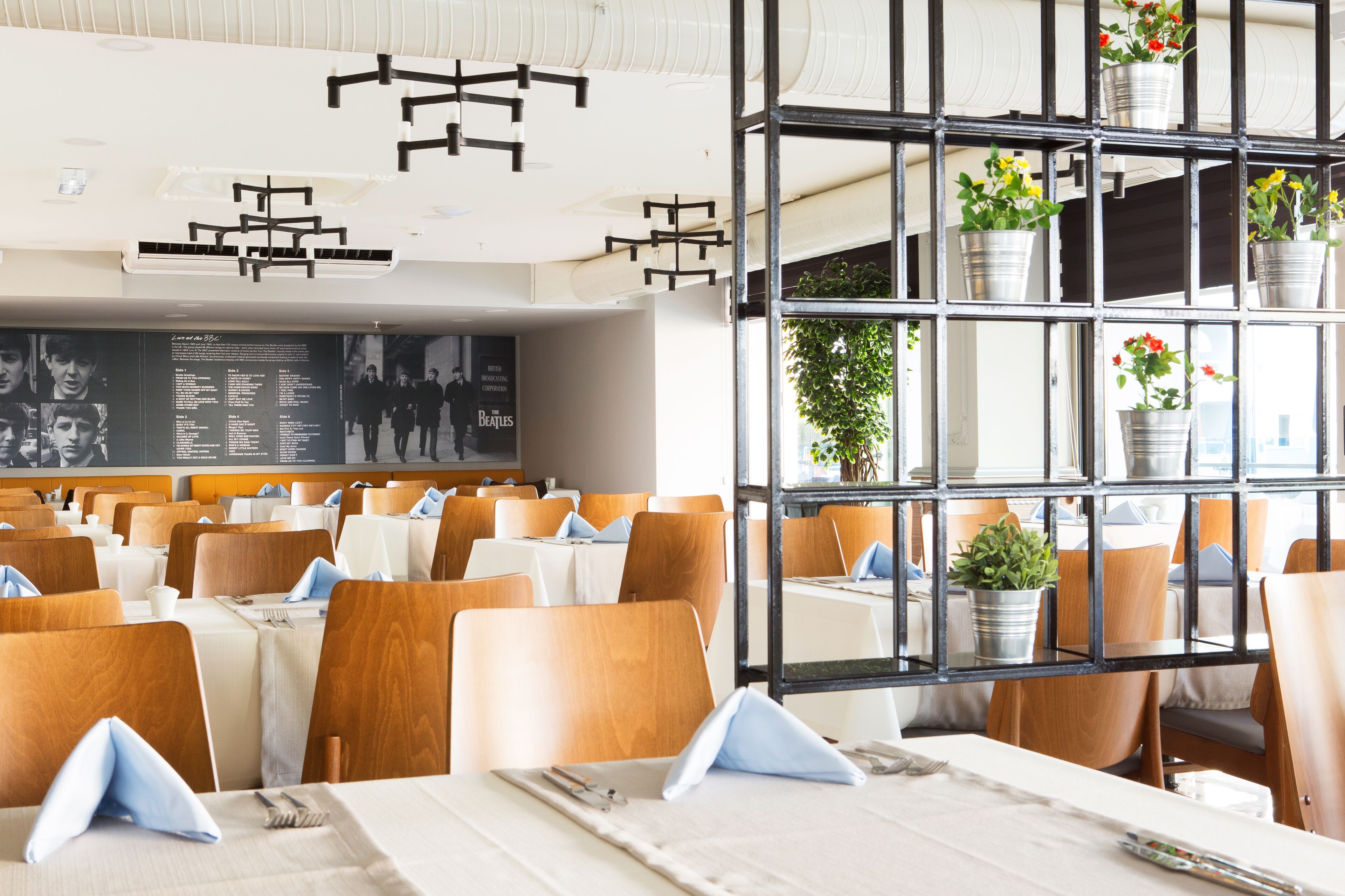 A restaurant with multiple tables, wooden chairs, and a black metal shelf with potted plants.