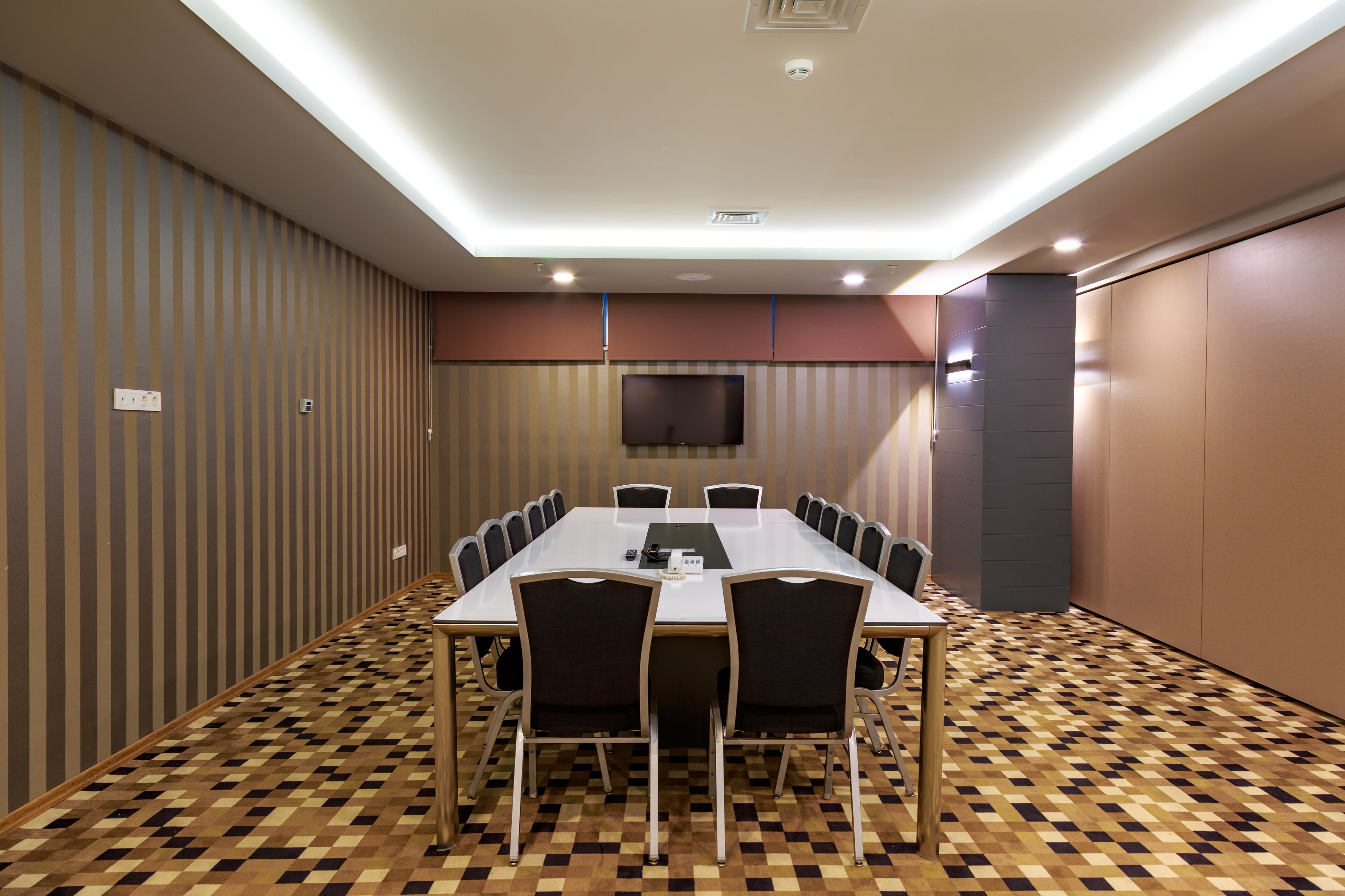 A conference room with a long table, chairs, a flat-screen TV, and patterned carpet.