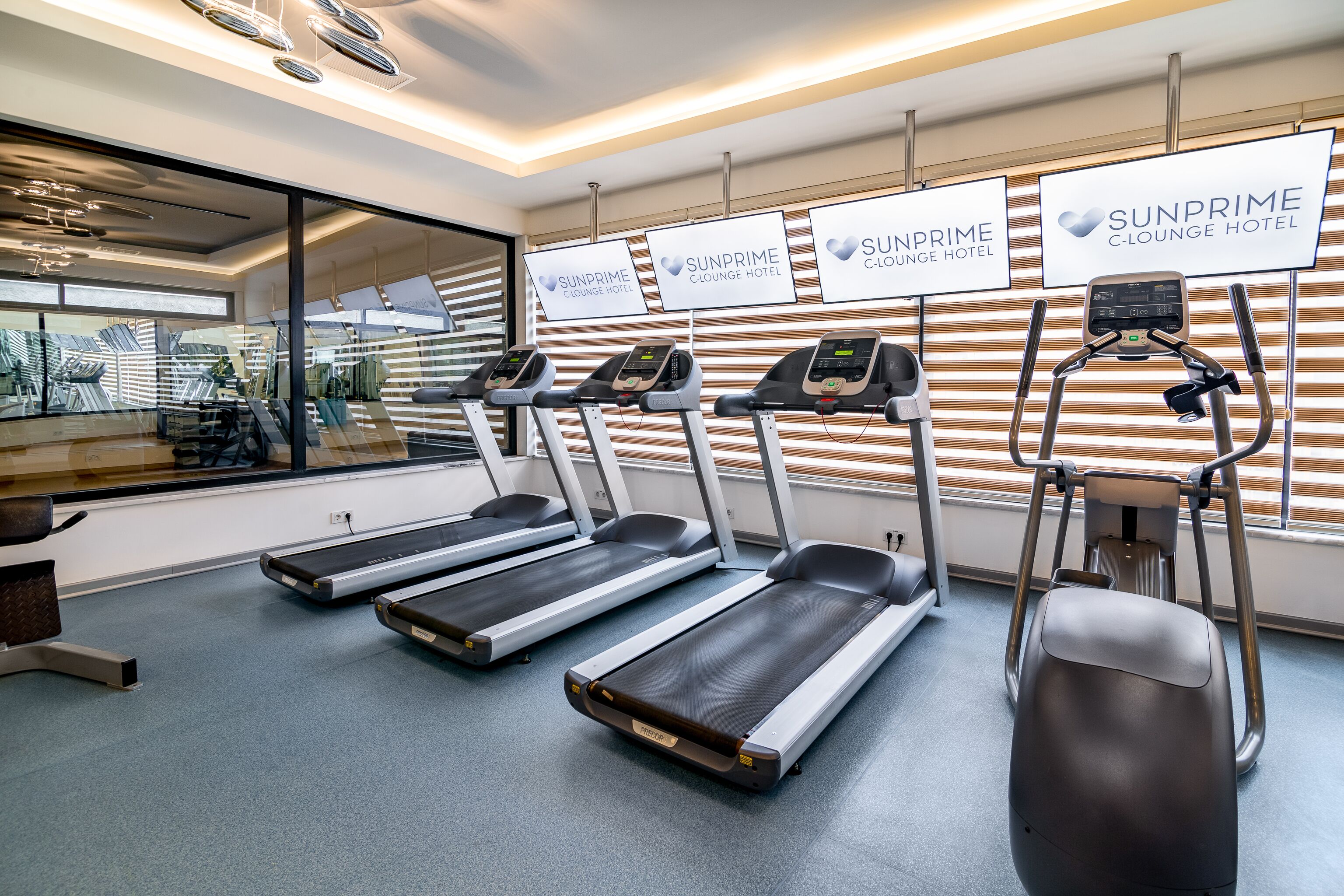 Et fitnessområde med løbebånd og trappemaskine, med skilte for SunPrime C-Lounge Hotel på væggen.