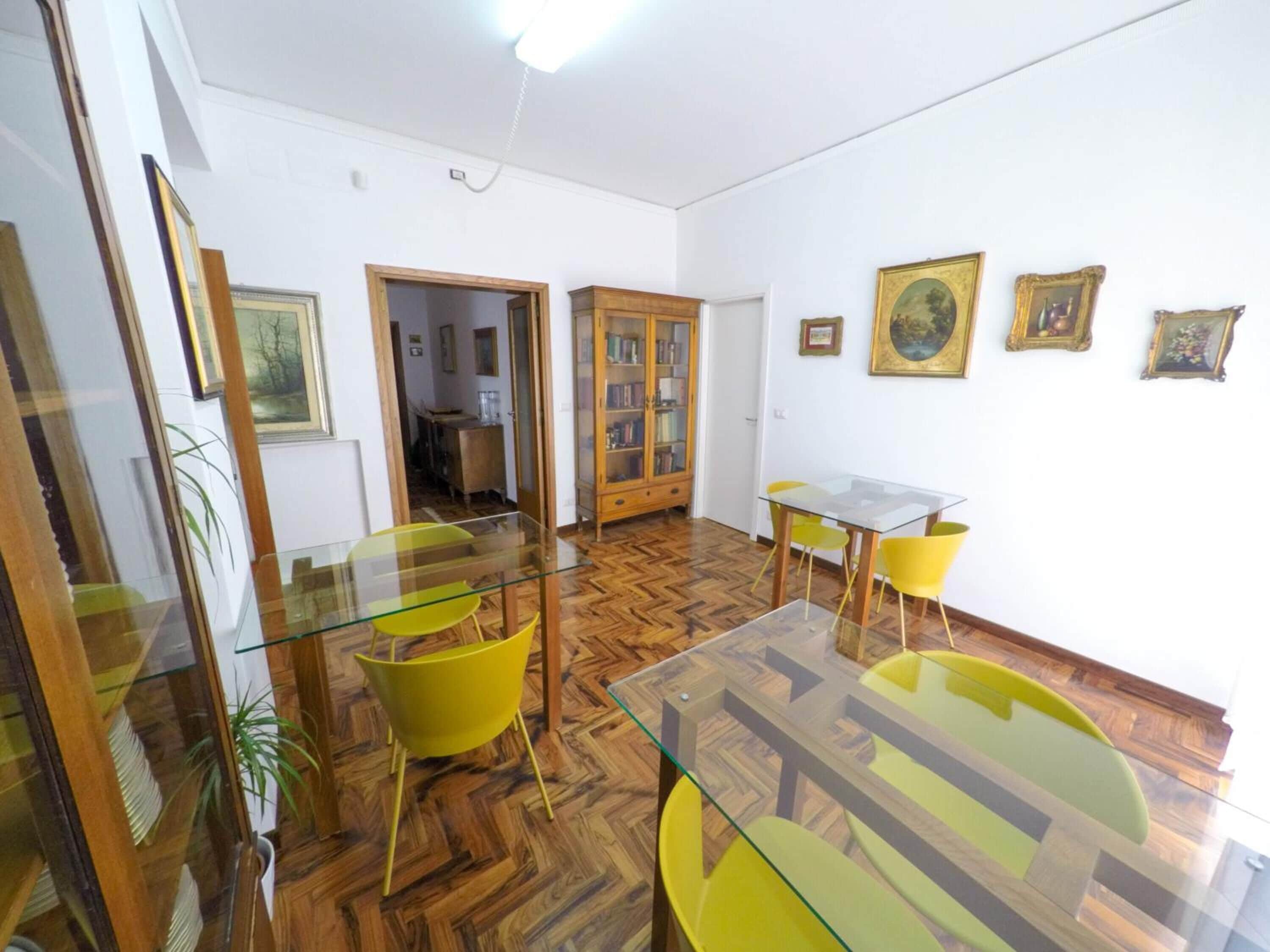 A dining area with a glass table and yellow chairs, a wooden cabinet with books, and a herringbone-patterned floor.