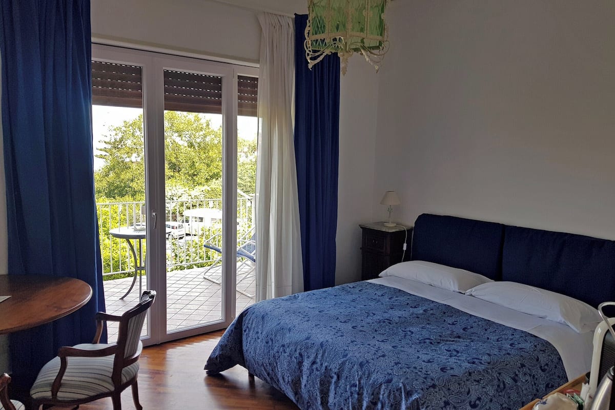 Et soveværelse med en seng, et lille bord og en stol. Der er en balkon med udsigt over grønt og en hvid bygning.