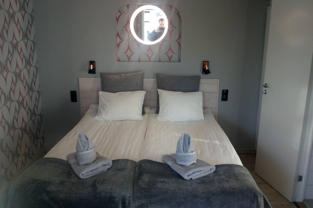 Una cama doble con dos almohadas, una mesita de noche con lámpara y un espejo instalado en la pared.