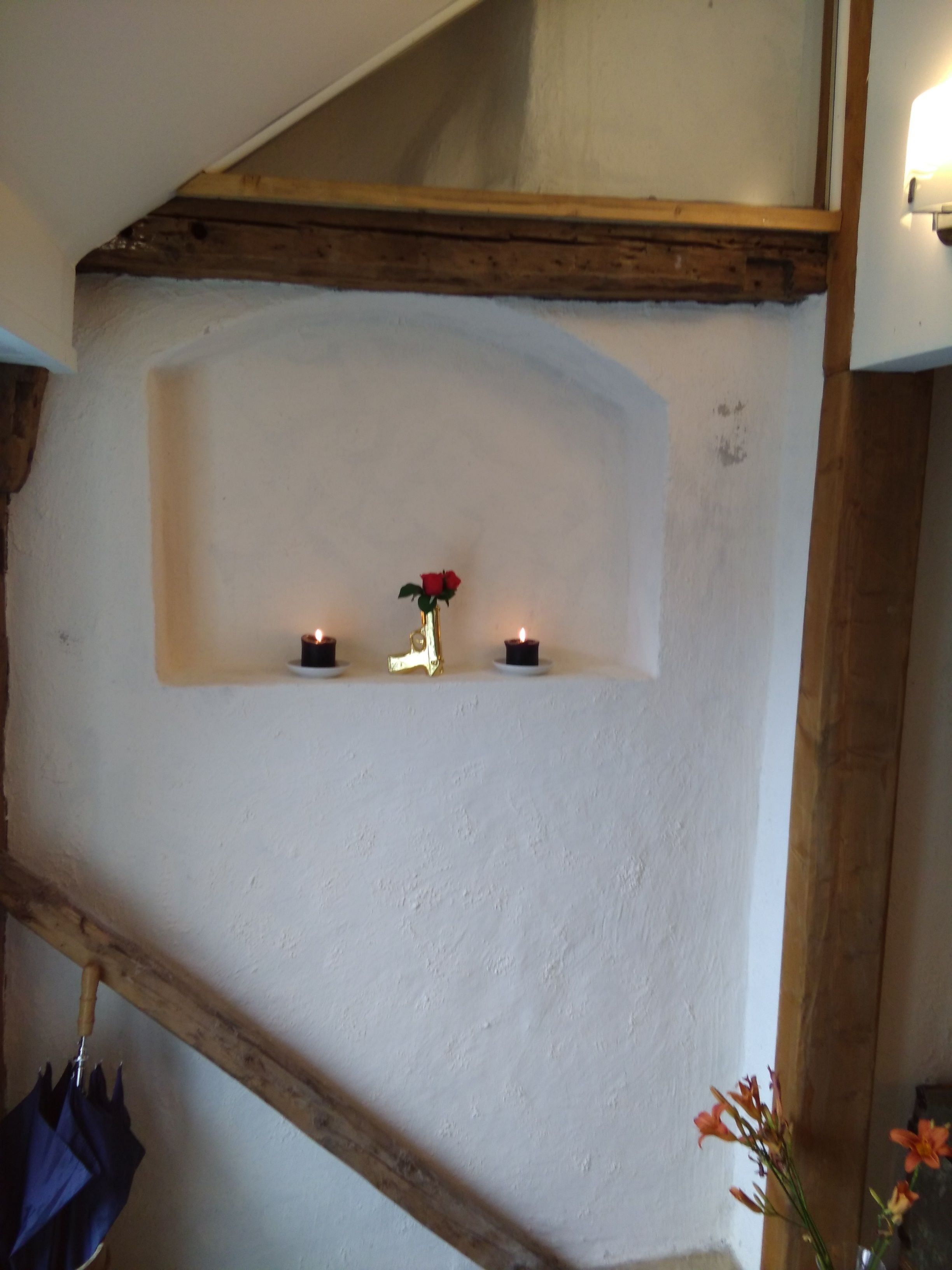 A room with a white textured wall, a wooden beam ceiling, and a shelf with candles and a vase with a red flower.
