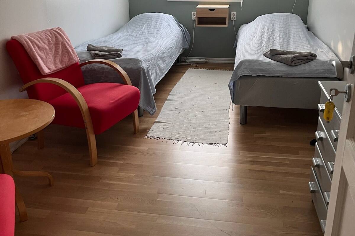 Una habitación pequeña y funcional con dos camas individuales, una silla roja, una mesita y una ventana con cortinas.