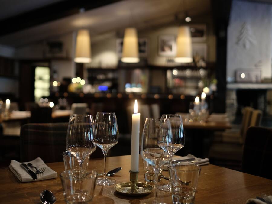 A dining table set with wine glasses, a candle, and cutlery, with a bar and ambient lighting in the background.
