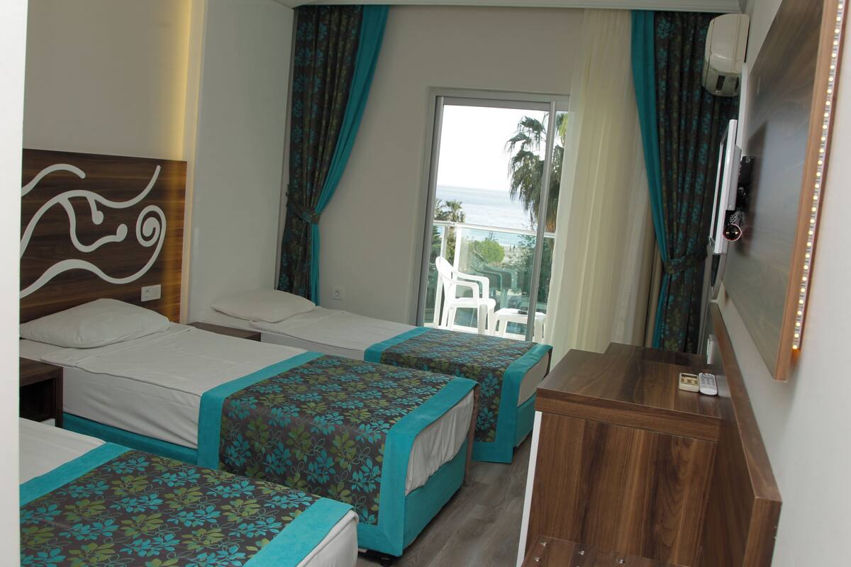 Ein Hotelzimmer mit zwei Betten, einem Holzkoffer, einem Fernseher und einem Balkon mit Blick aufs Meer.