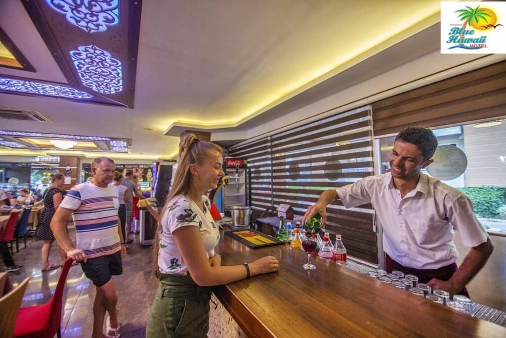 Un espace bar avec un barman qui prépare des boissons, une femme debout près du comptoir et un homme qui marche au fond.