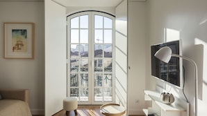 Une chambre moderne avec une grande fenêtre, un lit, un petit tabouret, une télévision et un bureau avec des étagères.