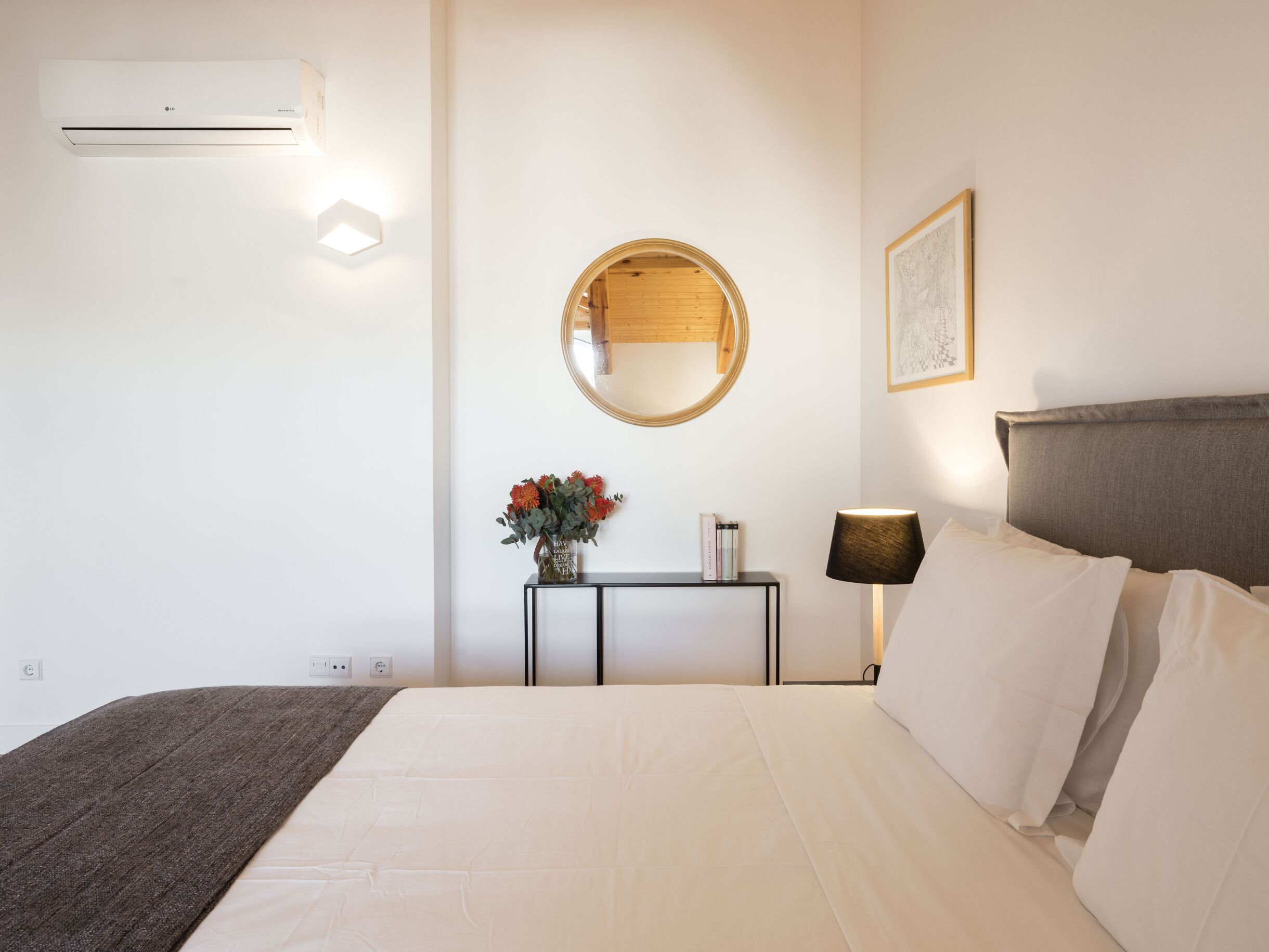 A hotel room with a bed, a bedside table with a lamp, a mirror, and a wall-mounted air conditioner.