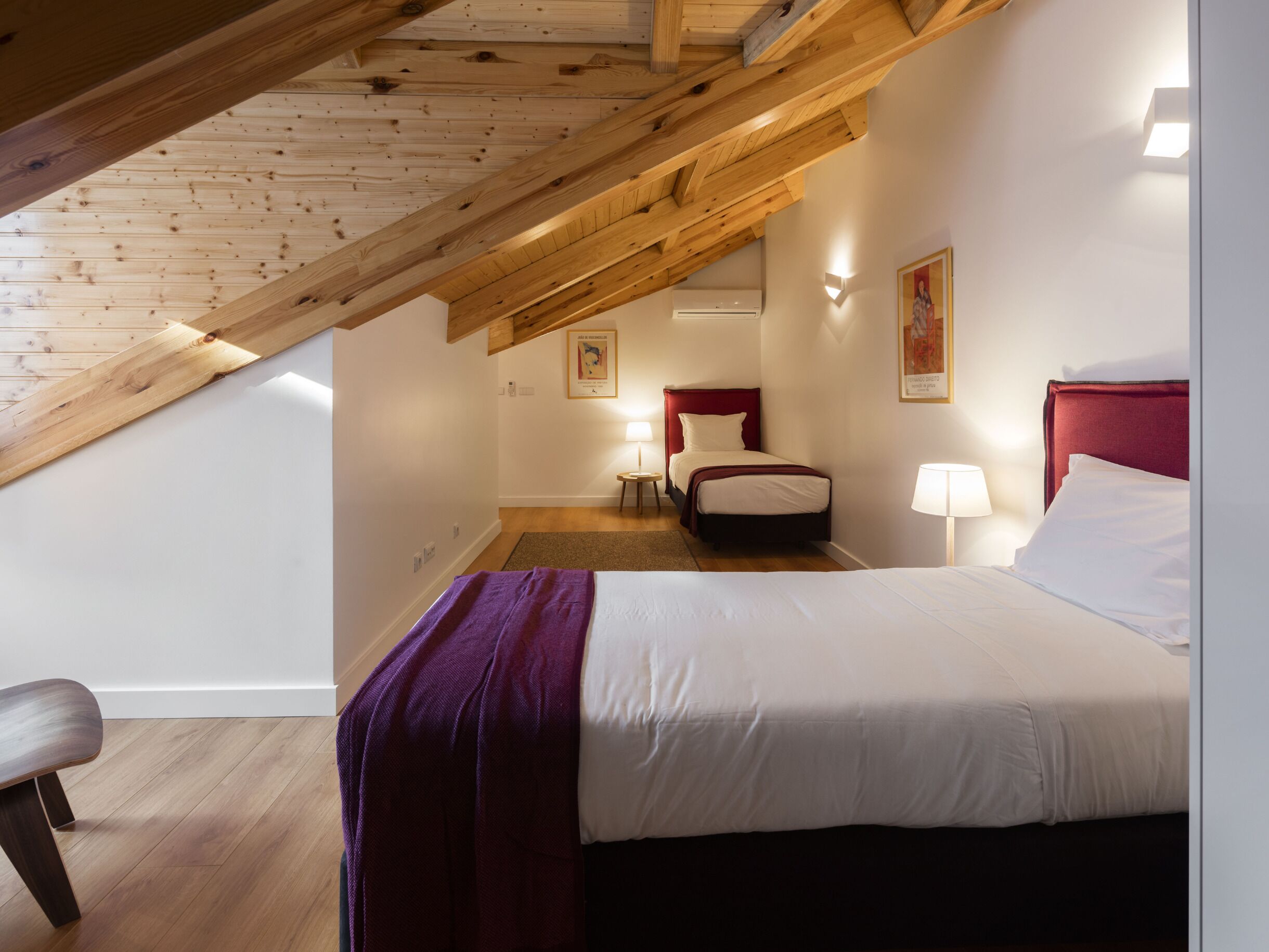 Ein Schlafzimmer mit schrägen Wänden, einem Bett mit weißer Bettwäsche, einem roten Kopfteil und einem kleinen Nachttisch mit Lampe.