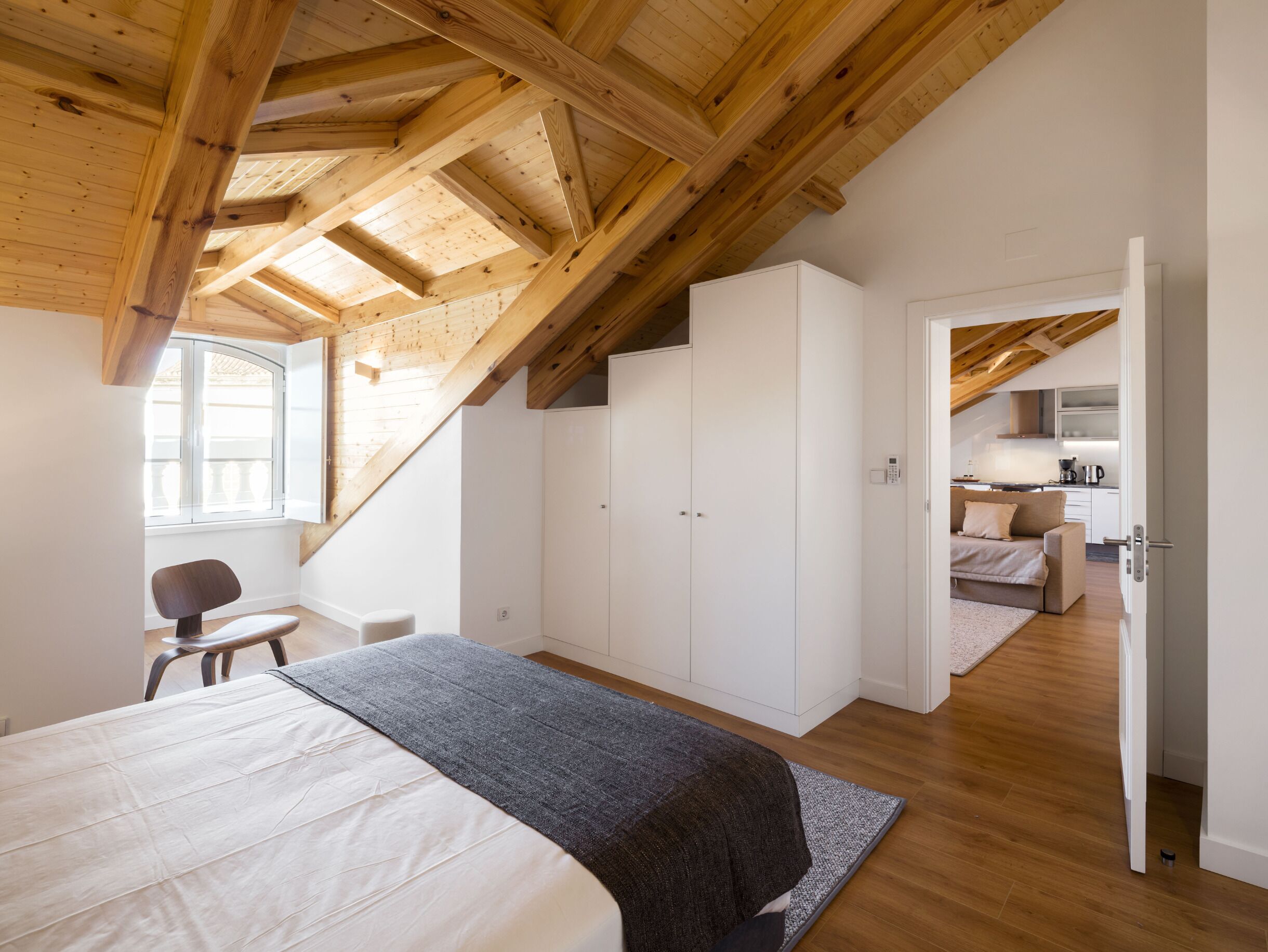 Een slaapkamer met een groot bed, een stoel en een kledingkast. Er is een dakraam en zicht op een keuken in de aangrenzende kamer.