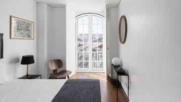Quarto moderno com uma cama grande, uma cadeira, uma escrivaninha, uma luminária e vista através de uma porta de vidro.