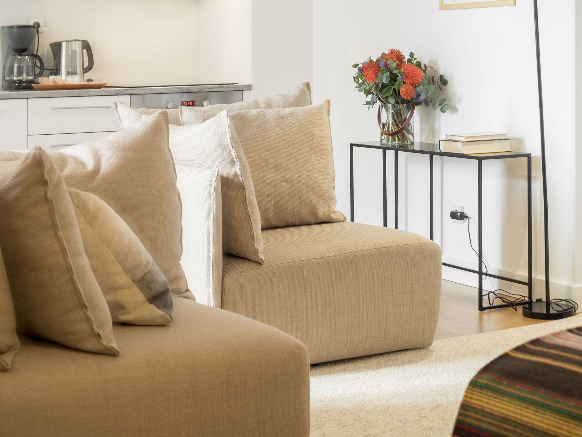 A modern living room with a beige sofa, a coffee maker, a vase with flowers, and a bookshelf.