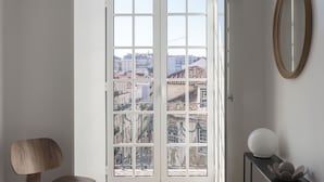 Une chambre avec une grande fenêtre, une chaise en bois, une table de chevet avec une lampe et un lit.