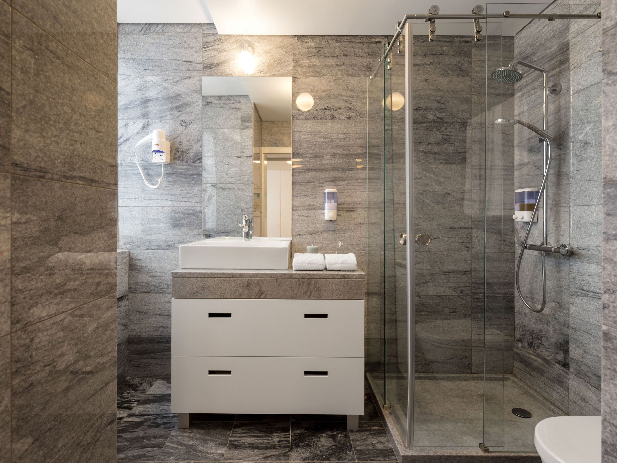 Une salle de bain moderne avec une douche à parois de verre, des murs en marbre et un meuble-lavabo blanc doté de tiroirs.