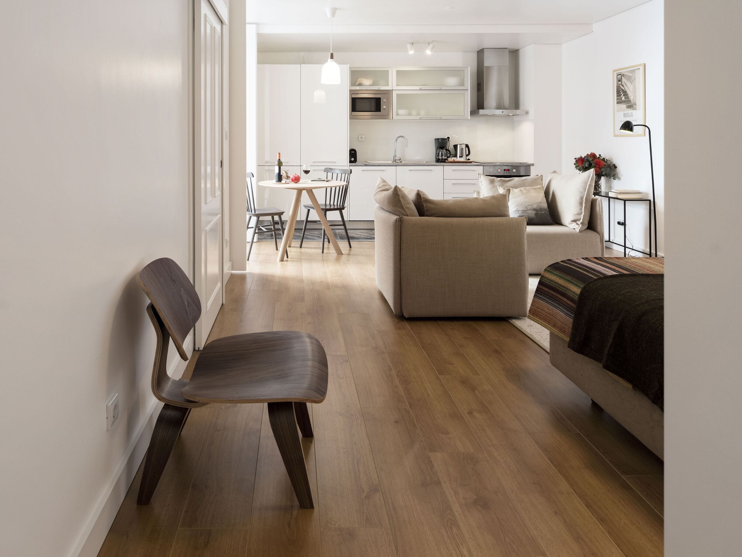 A modern living room with a wooden floor, a sofa, a dining table with chairs, and a kitchen area in the background.