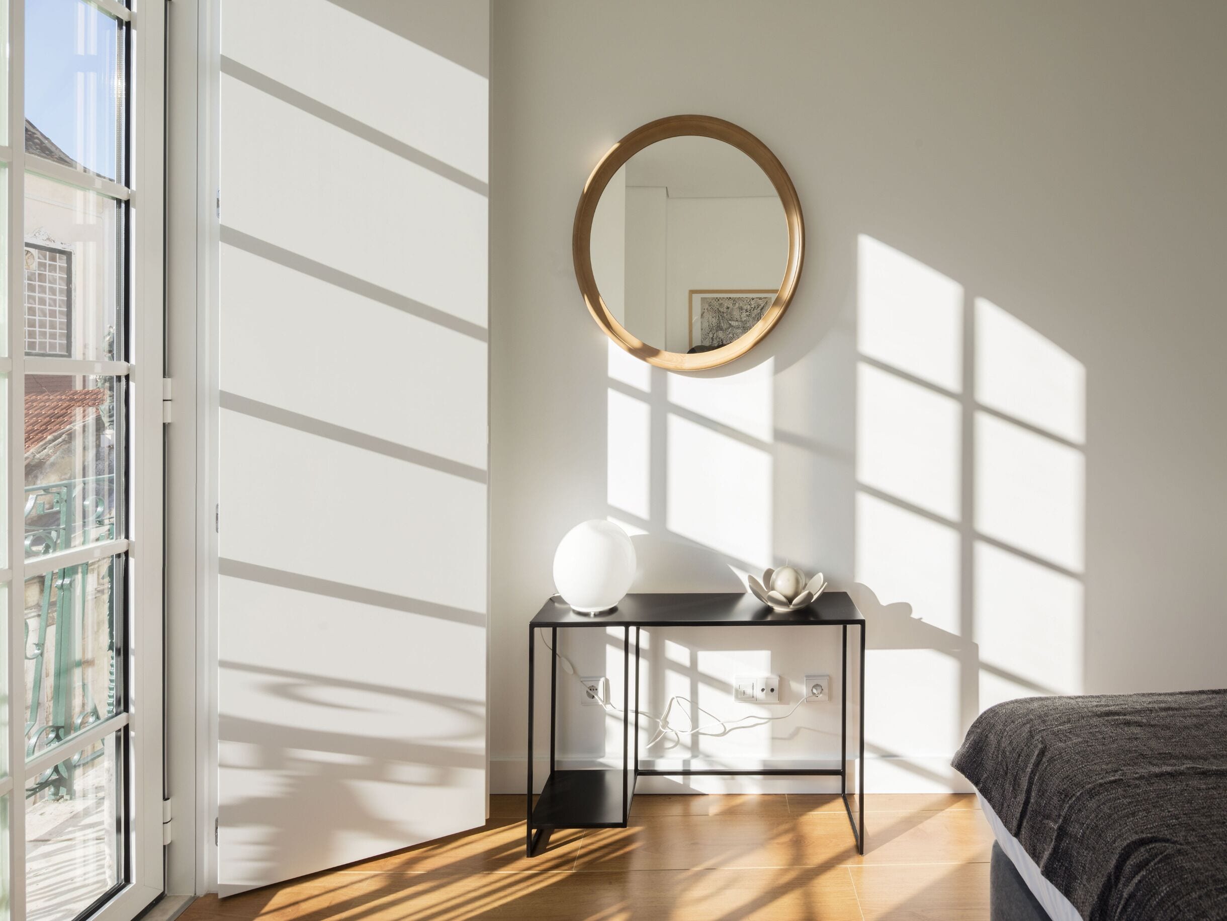 Quarto moderno com uma janela ampla, um espelho redondo, mesa de cabeceira com abajur e uma cama.
