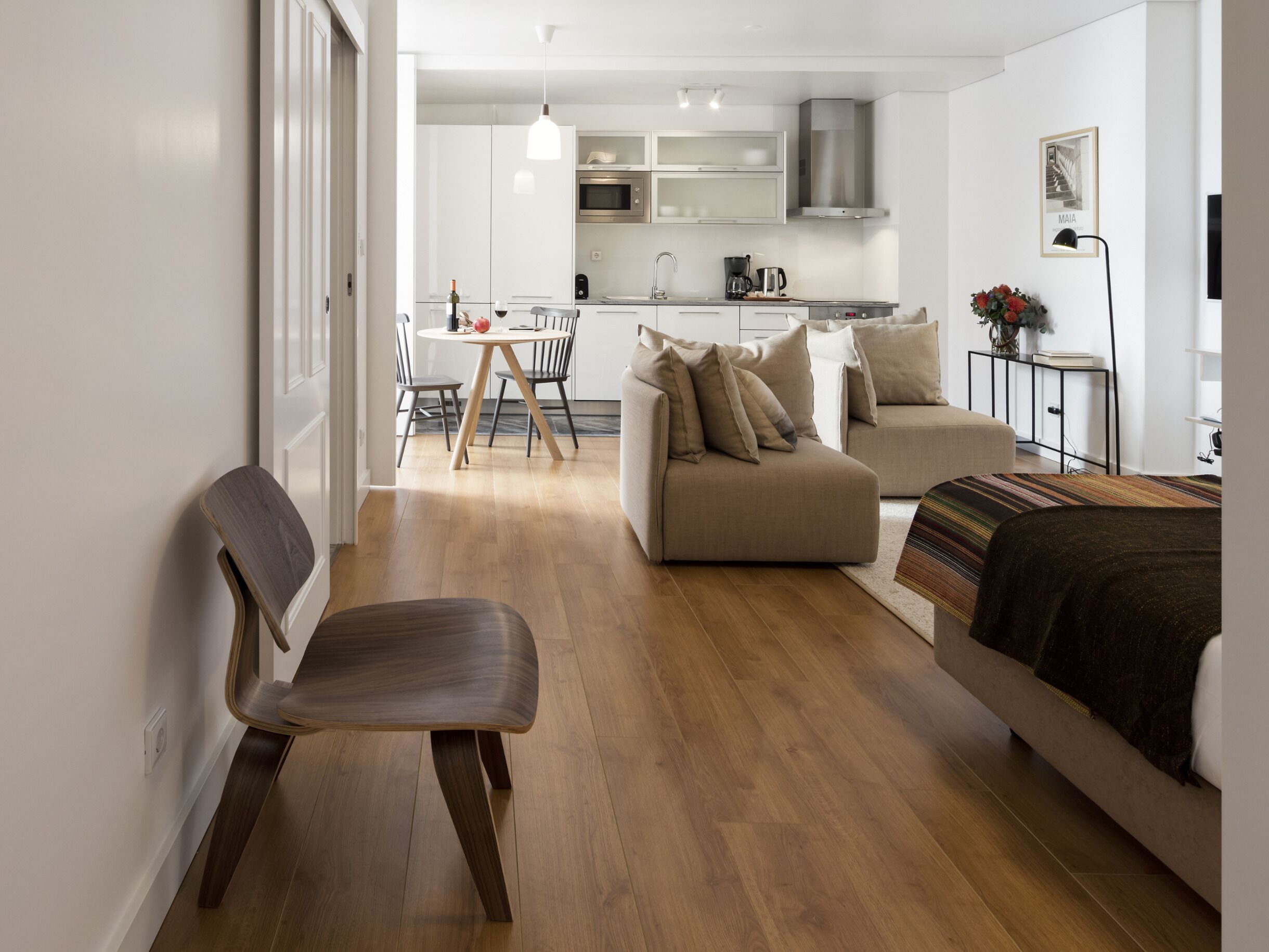 A modern living room with a sofa, a dining table with chairs, a kitchen area with cabinets and appliances, and a hallway leading to another room.
