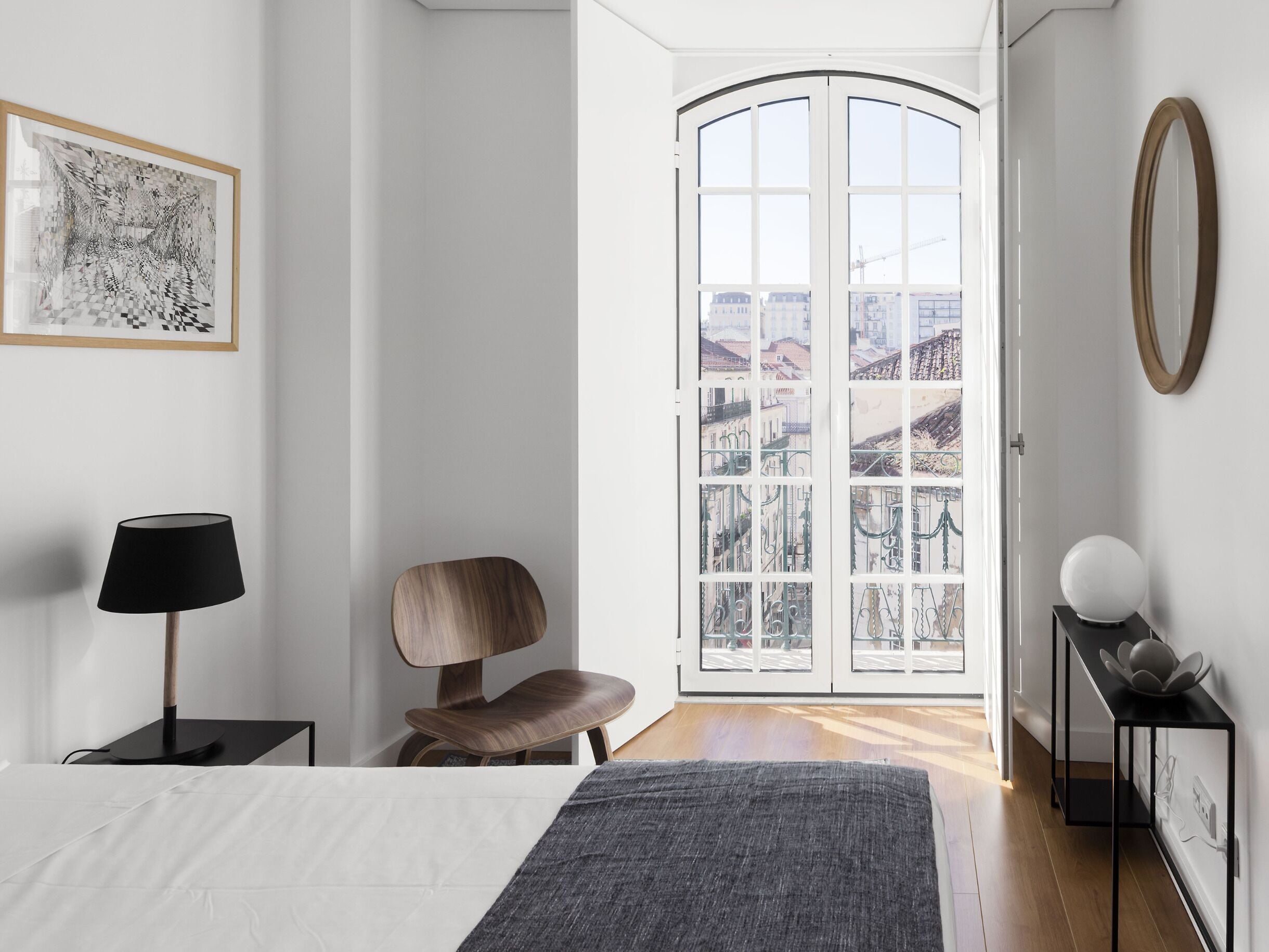 Un dormitorio moderno con una cama grande, una silla, un escritorio y vistas de edificios a través de un ventanal.