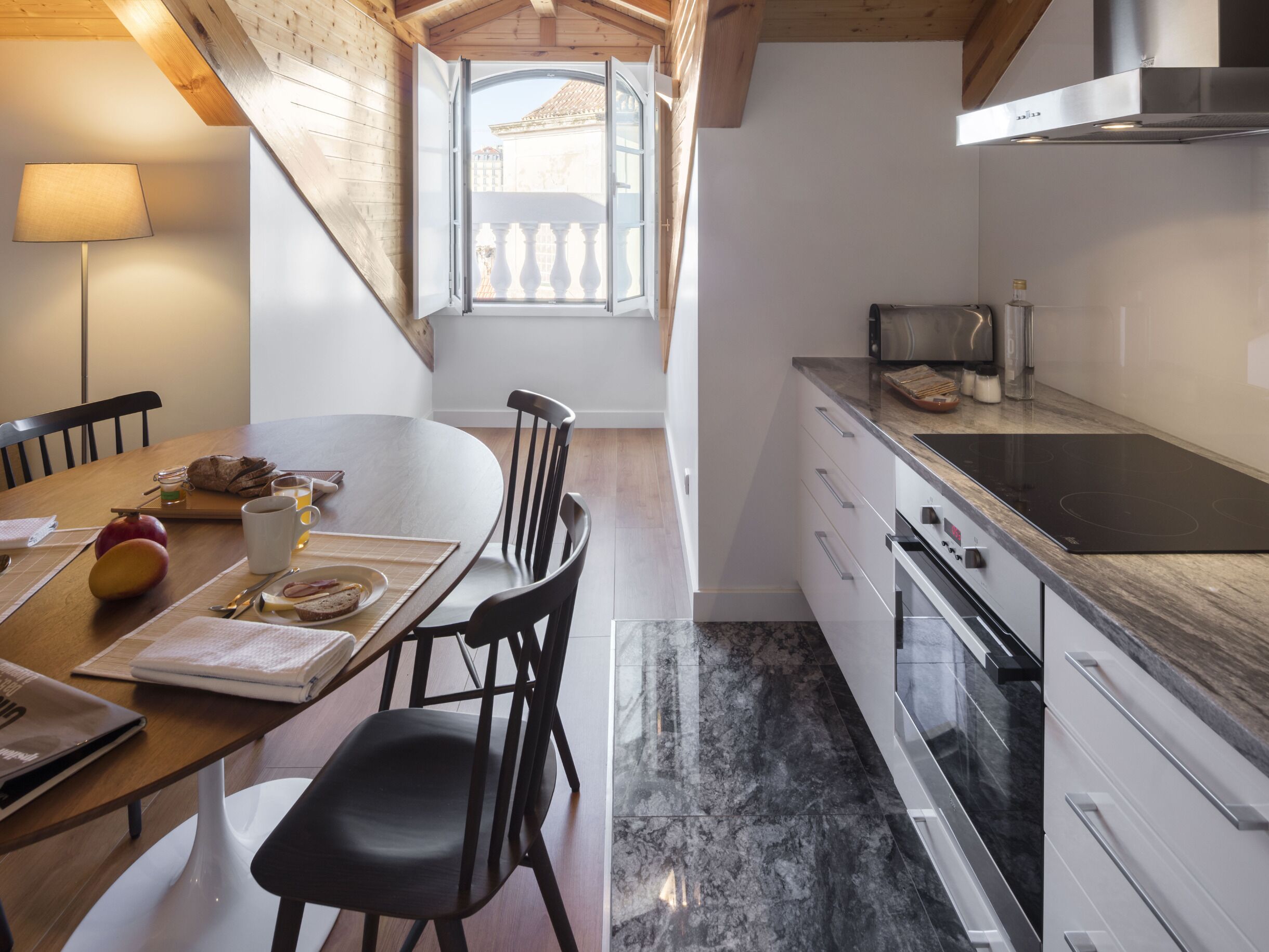 A modern kitchen with a dining area, featuring a wooden ceiling, a large window with a balcony, and a stainless steel range hood.
