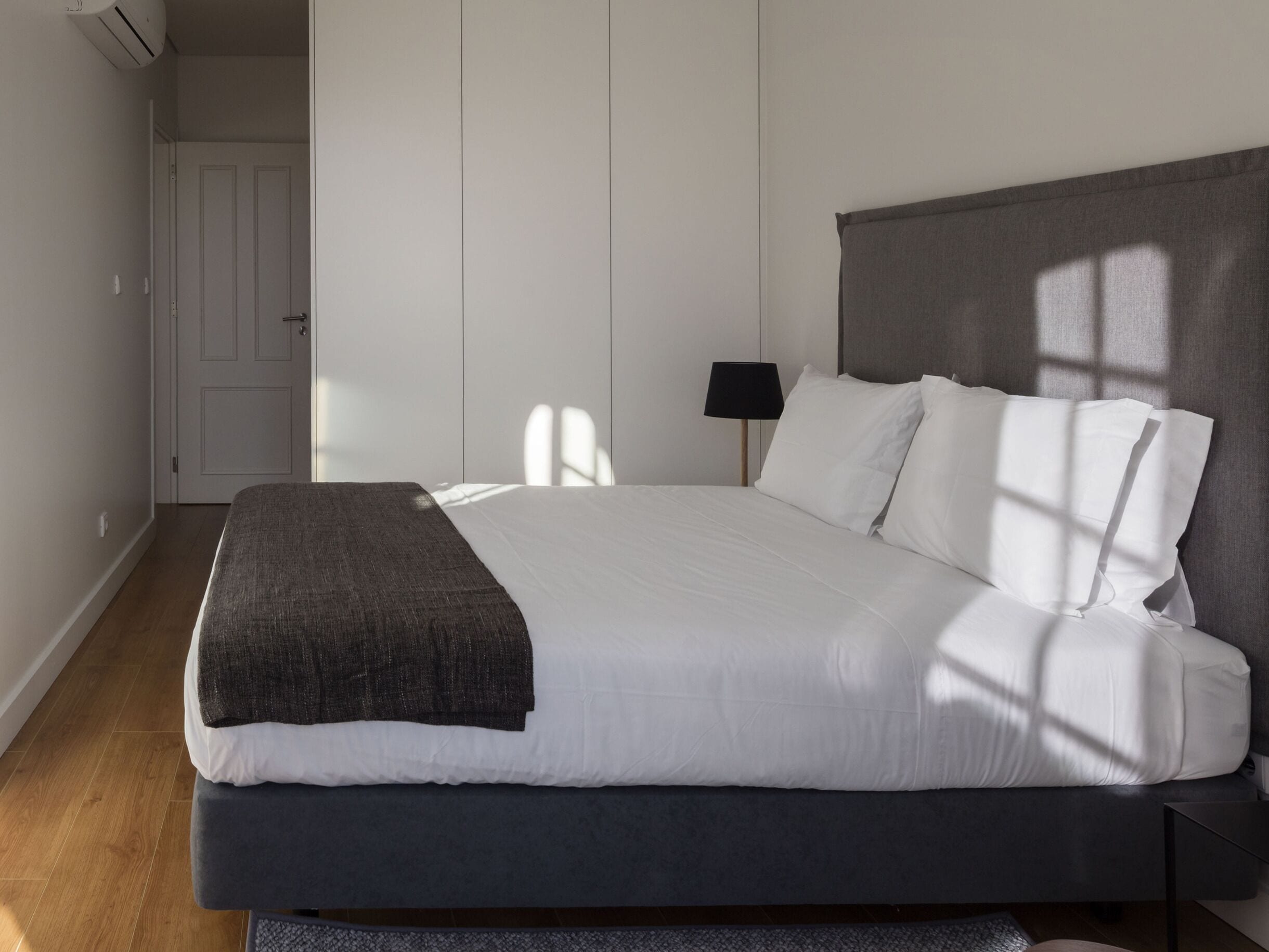 Una cama bien hecha con ropa de cama blanca y gris, un cabecero oscuro y una mesita de noche pequeña con una lámpara.