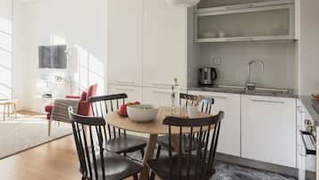 A modern kitchen with white cabinets, a stainless steel sink, and a built-in oven. There is a dining table with chairs and a bowl of fruit.