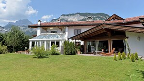 A two-story house with a large glass conservatory, a patio with outdoor furniture, and a mountain backdrop. - Landhaus Florian (Sankt Johann in Tirol)