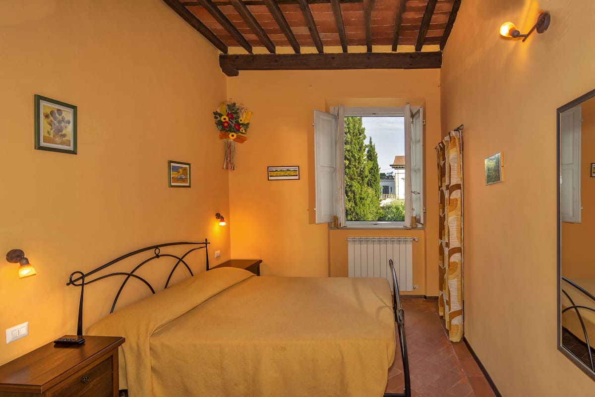 A bedroom with a bed, bedside lamps, a window with curtains, and a view of greenery outside.