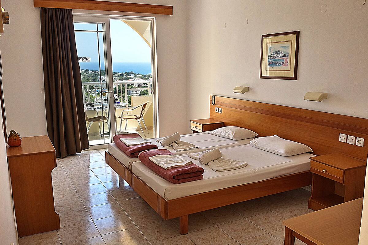 A hotel room with a bed, a wooden nightstand, a small table, and a view of the sea through the window.