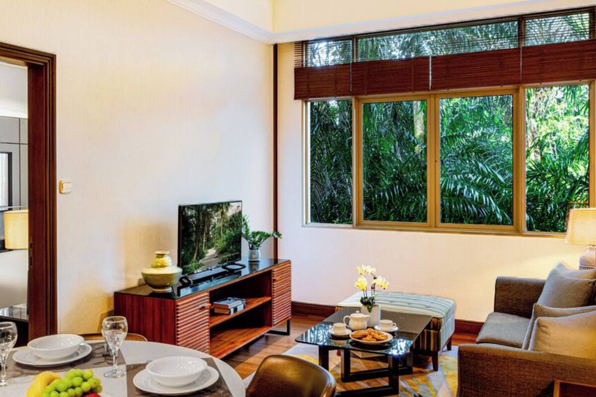 A hotel room with a bed, a TV, a dining area with a table set for two, and a window with a view of greenery.