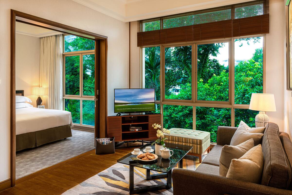 A hotel room with a bed, a sofa, a TV, and a coffee table with a view of greenery outside.