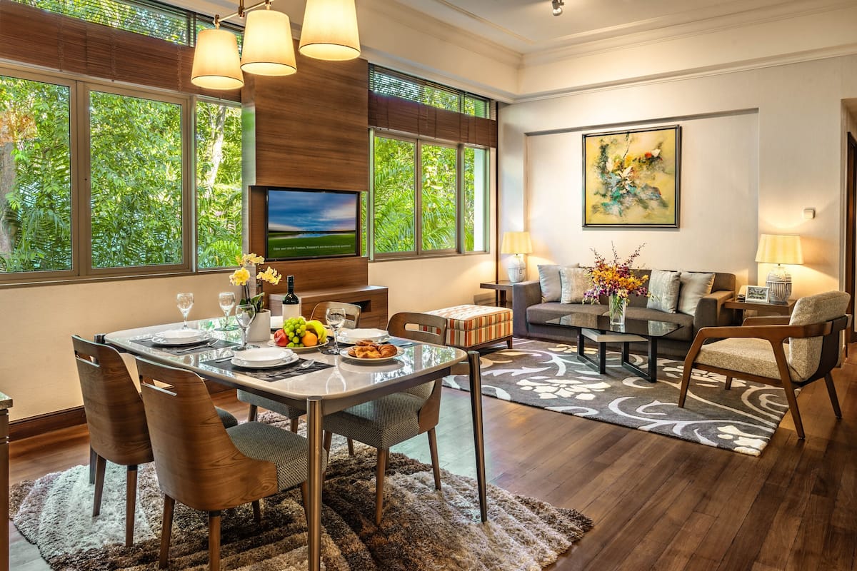 A modern dining area with a glass table, wooden chairs, and a view of lush greenery through large windows.