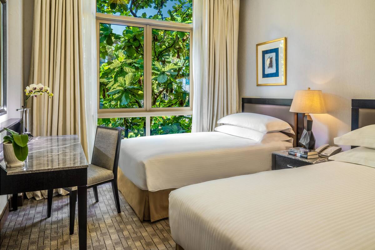 A hotel room with two beds, a desk with a chair, a lamp, and a window with curtains.