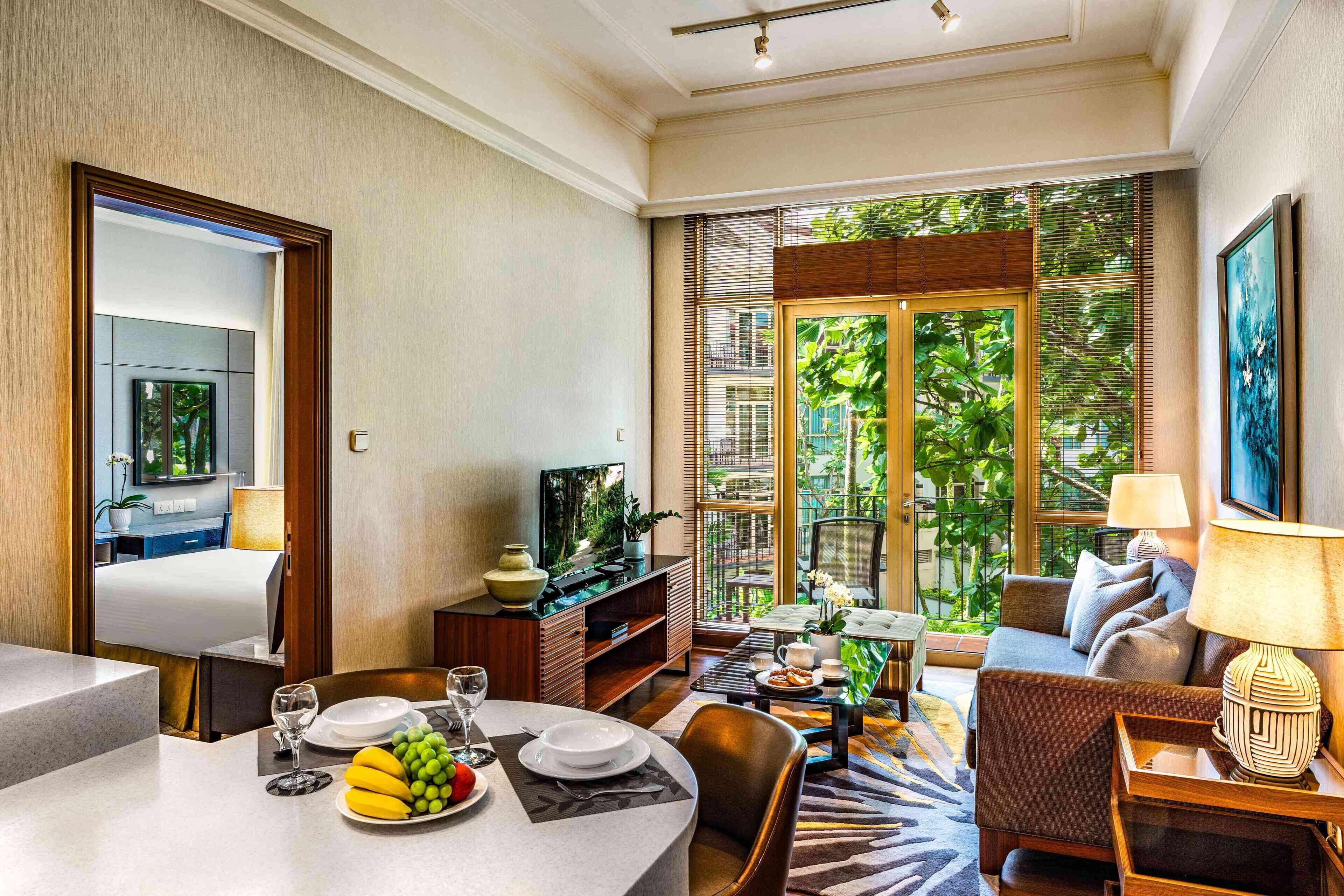 A modern hotel room with a dining area, a sofa, and a large window with a view of greenery.