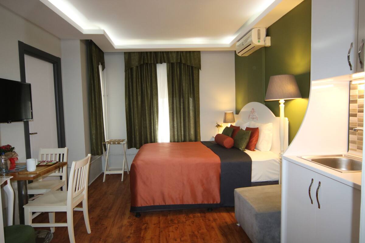 Habitación de hotel con cama, escritorio con silla, kitchenette pequeña y televisor montado en la pared.