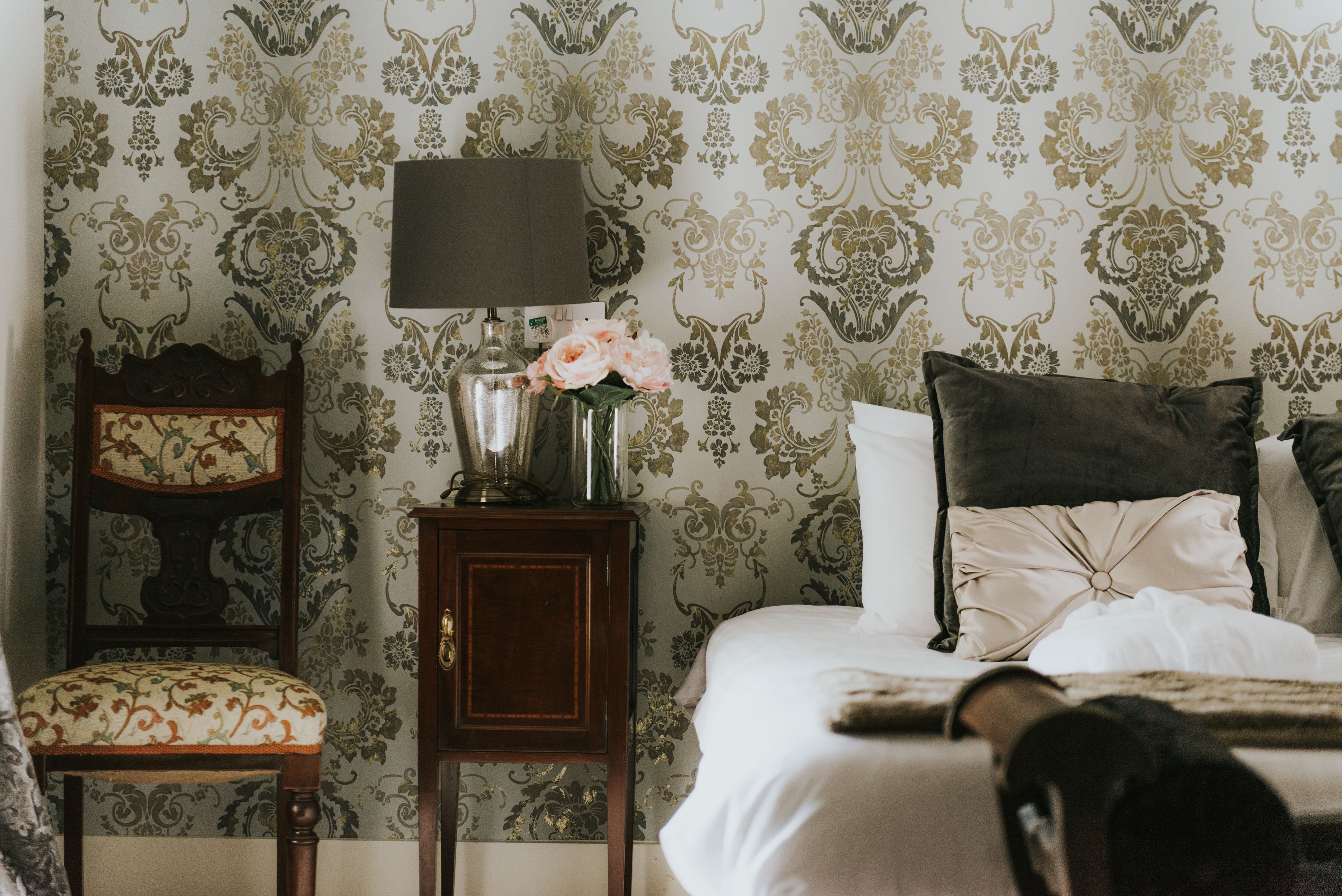 Un dormitorio con cama, mesita de noche con lámpara, una silla y un florero con flores.